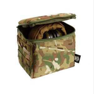 Könnte beinhalten: Eine quadratische Tasche mit Tarnmuster und Reißverschluss oben, die teilweise geöffnet ist, um einen beigefarbenen Innenraum und einen Blick auf ein dunkles Objekt freizugeben. Die Tasche hat eine grüne Einfassung und kleine Außentaschen. Ein schwarzes Etikett mit weißem Logo ist angebracht.