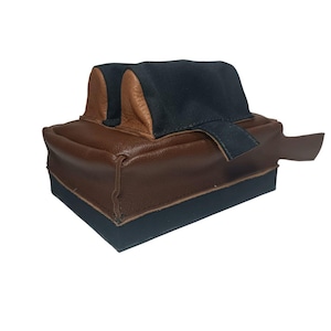 May include: A rectangular shooting rest featuring a brown leather base and sides, topped with two black fabric-covered supports. The supports have brown leather accents on the inner sides and a black fabric strap extending from the right side.