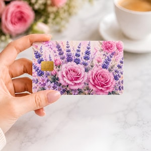 Puede incluir: Una tarjeta de cr&eacute;dito con dise&ntilde;o floral que presenta rosas rosas y lavanda p&uacute;rpura. La tarjeta tiene un chip dorado y se sostiene en una mano. En el fondo hay una taza de caf&eacute; y un ramo de flores.