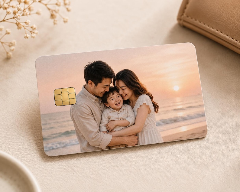 May include: A personalized credit card featuring a family portrait at the beach during sunset. The card has a gold-colored chip and is surrounded by a neutral-toned background with a wallet and decorative flowers.
