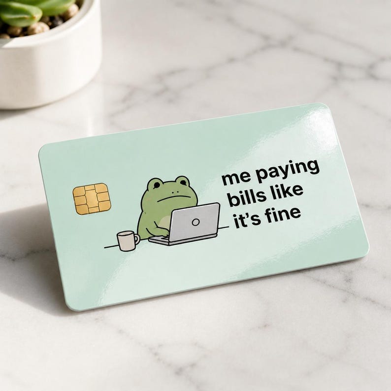 May include: A light green credit card with a cartoon frog using a laptop. The card has the text "me paying bills like it's fine". A small cup and a gold chip are also visible.
