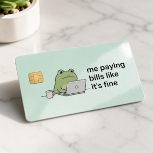 May include: A light green credit card with a cartoon frog using a laptop. The card has the text "me paying bills like it's fine". A small cup and a gold chip are also visible.