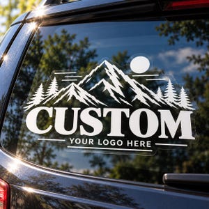 May include: A white vinyl decal applied to a car window featuring a mountain range, pine trees, and a sun. The text CUSTOM is printed in large bold letters, with the text YOUR LOGO HERE positioned below it, framed by horizontal lines.