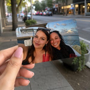 Op de afbeelding: Een gepersonaliseerde creditcard met een foto van twee lachende vrouwen op een boot. De kaart heeft een witte chip en wordt vastgehouden tegen een vage buitenachtergrond.