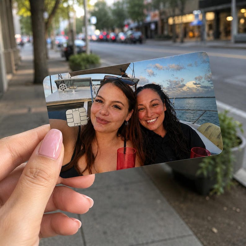 Op de afbeelding: Een gepersonaliseerde creditcard met een foto van twee lachende vrouwen op een boot. De kaart heeft een witte chip en wordt vastgehouden tegen een vage buitenachtergrond.