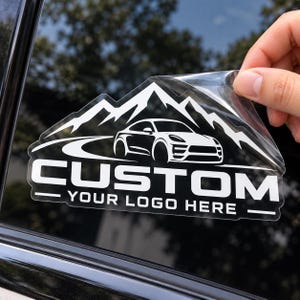 May include: A white vinyl decal for a car window featuring a graphic of a sports car driving on a road in front of mountain peaks. The text CUSTOM is printed in a bold, sans-serif font above the smaller text YOUR LOGO HERE, all on a clear backing.