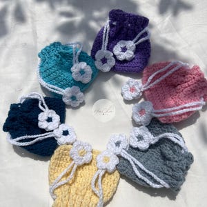 Könnte beinhalten: Eine Sammlung handgefertigter Häkeltaschen in verschiedenen Farben, darunter Blau, Lila, Rosa, Gelb und Grau. Jede Tasche hat einen Kordelzugverschluss und ist mit weißen Häkelblumen verziert. Die Taschen sind kreisförmig angeordnet.