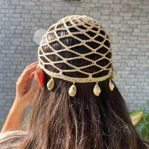 Könnte beinhalten: Eine beige Häkelkopfbedeckung mit Rautenmuster, verziert mit hängenden Muscheln. Eine rote Feder ist seitlich sichtbar. Die Kopfbedeckung wird von einer Person mit langen, dunkelbraunen Haaren getragen.