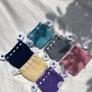 Può includere: Una collezione di sette piccoli sacchetti all'uncinetto in vari colori: blu navy, giallo, viola, rosa, grigio e azzurro. Ogni sacchetto ha un cordoncino bianco e un piccolo fiore bianco all'uncinetto. I sacchetti sono disposti su una superficie bianca.