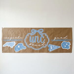 Könnte beinhalten: Ein braunes Banner mit hellblauem Text und Grafiken. Das Banner zeigt "congrats grad" mit "unc board" in der Mitte. Es gibt auch mehrere hellblaue Grafiken eines Hutes, einer Wimpel und des Bundesstaates North Carolina.