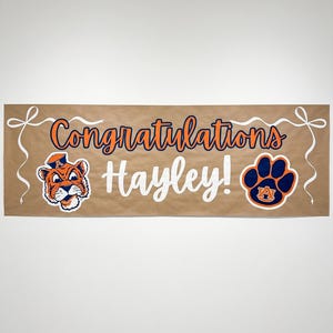 May include: A brown banner with the words "Congratulations Hayley!" in orange and white. It features a tiger mascot and a paw print logo, both in orange and blue. White ribbon accents are at the top corners.