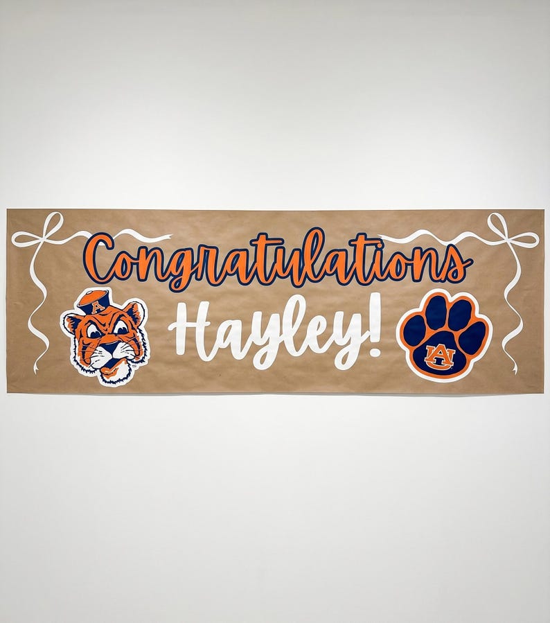 May include: A brown banner with the words "Congratulations Hayley!" in orange and white. It features a tiger mascot and a paw print logo, both in orange and blue. White ribbon accents are at the top corners.