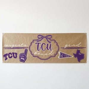 May include: A brown banner with purple text and graphics. The banner says "congrats grad" and features the letters "TCU" in a decorative font. Additional graphics include a foam finger, a pennant, and a Texas outline.