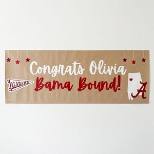 May include: A banner with the words "Congrats Olivia Bama Bound!" in white and red lettering. The banner includes a graphic of the state of Alabama and a pennant with the word "Alabama". Red stars are strung across the top.