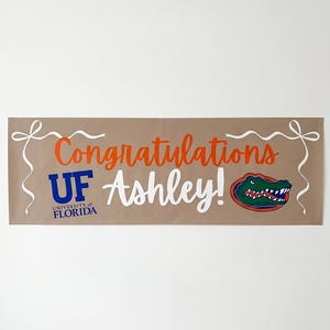 May include: A tan banner with the words "Congratulations Ashley!" in orange and white script. The University of Florida logo is in blue, and a Gator logo is on the right. White ribbon-like accents are on each end.
