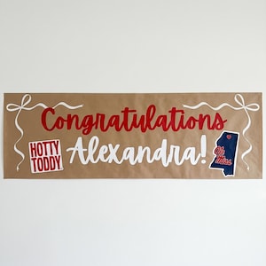 May include: A brown banner with the words "Congratulations Alexandra!" in red and white script. The banner includes a "Hotty Toddy" logo and a Mississippi state outline with "Ole Miss" text.