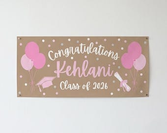 May include: A graduation banner in shades of brown, pink, and white. The banner reads "Congratulations Kehlani Class of 2026" with pink balloons, a graduation cap, and a diploma illustration. The banner is rectangular and mounted on a wall.