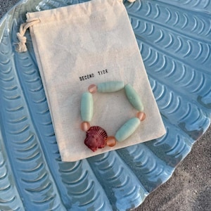 Op de afbeelding: Een armband met zeegroene en perzik kleurige kralen en een rode schelpenbedel ligt op een lichtblauwe schelpvormige schaal. Een kleine canvas tas met de tekst "SECOND TIDE" is ook aanwezig.