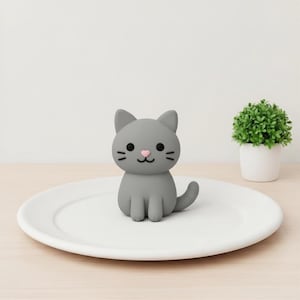 Puede incluir: Una pequeña figura de gato gris y lisa con cuerpo redondeado, orejas puntiagudas y cola curva. El gato tiene ojos negros circulares, tres bigotes negros en cada mejilla y una pequeña nariz rosa en forma de corazón, colocada sobre un plato blanco.