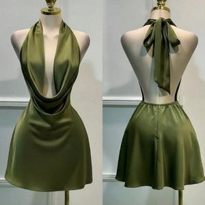 Puede incluir: Un vestido corto de satén color verde oliva con un escote drapeado profundo y diseño halter. La vista posterior muestra un cierre de lazo en el cuello, espalda descubierta y una falda acampanada. La prenda se exhibe en un maniquí.