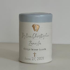 A blue glaze and white porcelain keepsake infant urn. It features a baby footprint, the custom name, a phrase, a gold cross, and the date.