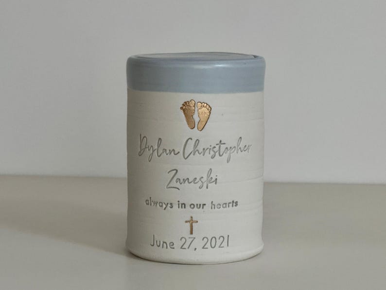 A blue glaze and white porcelain keepsake infant urn. It features a baby footprint, the custom name, a phrase, a gold cross, and the date.