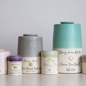 A collection of ceramic urns in various sizes and colors, including pink, gray, and teal. Some urns feature text such as names, dates, and the phrase "Always in our Hearts." The urns have a minimalist modern timeless design.