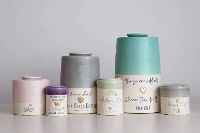 A collection of ceramic urns in various sizes and colors, including pink, gray, and teal. Some urns feature text such as names, dates, and the phrase "Always in our Hearts." The urns have a minimalist modern timeless design.