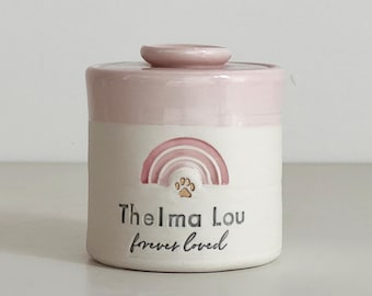 Op de afbeelding: Een kleine, cilindrische keramische pot met een deksel, beschilderd in wit en lichtroze. De pot heeft een regenboog- en pootafdrukontwerp, met de tekst "Thelma Lou forever loved" in zwart schrift. De pot is waarschijnlijk voor huisdierandenken.