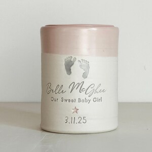 A pink glaze and white porcelain keepsake infant urn. It features a custom footprint, the custom name, a phrase, a star stamp, and the date.