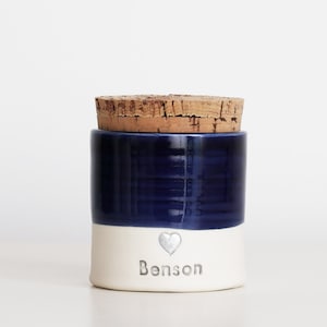 a custom cremation urn by vitrifiedstudio with personalized text and choice of stamps. color of urn can also be selected. urns come in various sizes. contact vitrifiedstudio via Etsy and we can assist you with the custom design of the urn. Image shows a deep blue glaze color on the top portion of the urn, with unglazed white clay on the lower portion. There is a cork lid. A small heart in silver with the name below.