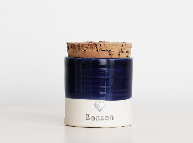 a custom cremation urn by vitrifiedstudio with personalized text and choice of stamps. color of urn can also be selected. urns come in various sizes. contact vitrifiedstudio via Etsy and we can assist you with the custom design of the urn. Image shows a deep blue glaze color on the top portion of the urn, with unglazed white clay on the lower portion. There is a cork lid. A small heart in silver with the name below.