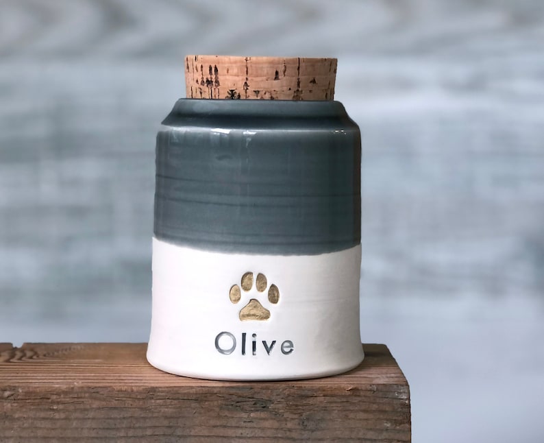 a custom cremation urn by vitrifiedstudio with personalized text and choice of stamps. color of urn can also be selected. urns come in various sizes. contact vitrifiedstudio via Etsy and we can assist you with the custom design of the urn. Image shows a collared shape urn with cork lid. It has a grey glazed upper section, and white unglazed clay lower portion. There is a gold paw stamp with name below