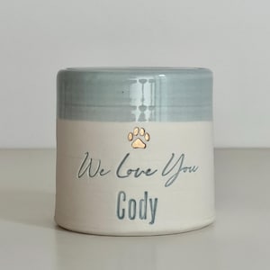 A handmade custom urn with a light blue lid and upper section, and a white lower section made of porcelain clay. The beautiful custom urn features a gold paw design, a custom phrase in cursive and a name below