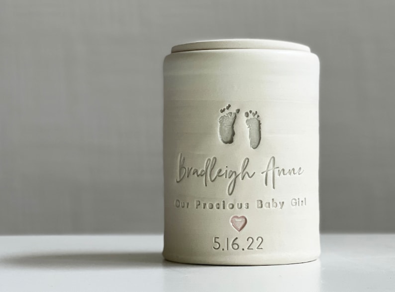 a custom cremation urn with personalized text and choice of stamps. color of urn can also be selected. urns come in various sizes. contact vitrifiedstudio via Etsy and we can assist you with the custom design of the urn. a custom footprint stamp can be made using your ink print image. Image shows medium size infant urn glazed in satin white with custom footprint stamp, pink heart stamp, and text.