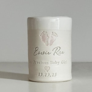 A small tall white glaze and white porcelain  keepsake infant urn. It features a custom footprint, the custom name, a phrase, a heart, and the date.