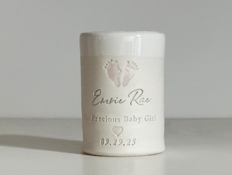 A small tall white glaze and white porcelain  keepsake infant urn. It features a custom footprint, the custom name, a phrase, a heart, and the date.