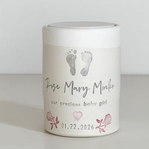 A pink glaze and white porcelain keepsake infant urn. It features a custom footprint, the custom name, a phrase, a heart, and the date with two rose stamps.