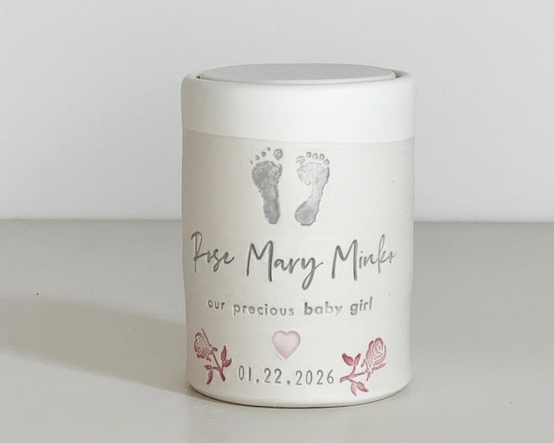 A pink glaze and white porcelain keepsake infant urn. It features a custom footprint, the custom name, a phrase, a heart, and the date with two rose stamps.