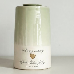 a custom cremation urn by vitrifiedstudio with personalized text and choice of stamps. color of urn can also be selected. urns come in various sizes. contact vitrifiedstudio via Etsy and we can assist you with the custom design of the urn. Image shows a two toned sage green glazed urn with a lower portion in white. There is a custom phrase in cursive over a gold heart over a name in cursive over dates.