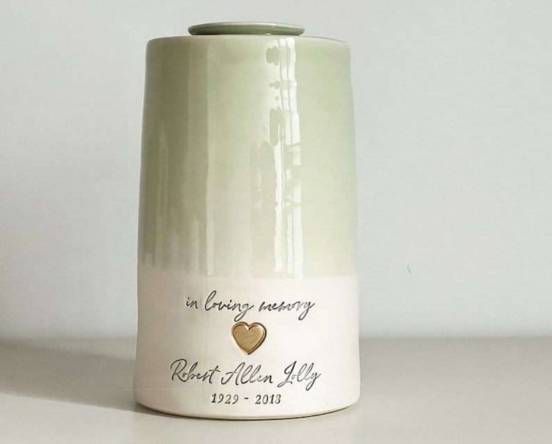 a custom cremation urn by vitrifiedstudio with personalized text and choice of stamps. color of urn can also be selected. urns come in various sizes. contact vitrifiedstudio via Etsy and we can assist you with the custom design of the urn. Image shows a two toned sage green glazed urn with a lower portion in white. There is a custom phrase in cursive over a gold heart over a name in cursive over dates.