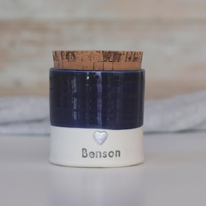 a custom cremation urn by vitrifiedstudio with personalized text and choice of stamps. color of urn can also be selected. urns come in various sizes. contact vitrifiedstudio via Etsy and we can assist you with the custom design of the urn. Image shows a deep blue glaze color on the top portion of the urn, with unglazed white clay on the lower portion. There is a cork lid. A small heart in silver with the name below.