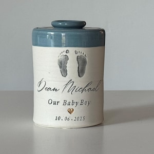 A small tall blue glaze and white porcelain  keepsake infant urn. It features a custom footprint, the custom name, a phrase, a gold heart, and the date.
