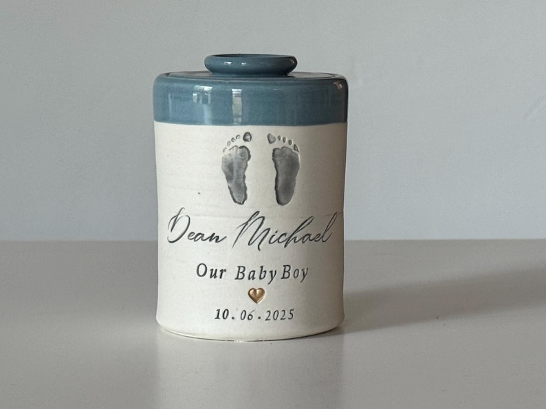 A small tall blue glaze and white porcelain  keepsake infant urn. It features a custom footprint, the custom name, a phrase, a gold heart, and the date.