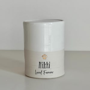A handmade custom urn with a white lid and upper section, and a white lower section made of porcelain clay. The beautiful custom urn features a gold paw design, name and a a custom phrase in cursive below