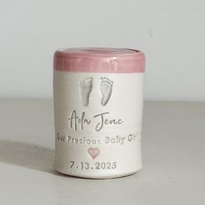 A small tall pink glaze and white porcelain keepsake infant urn. It features a custom footprint, the custom name, a phrase, a heart, and the date.
