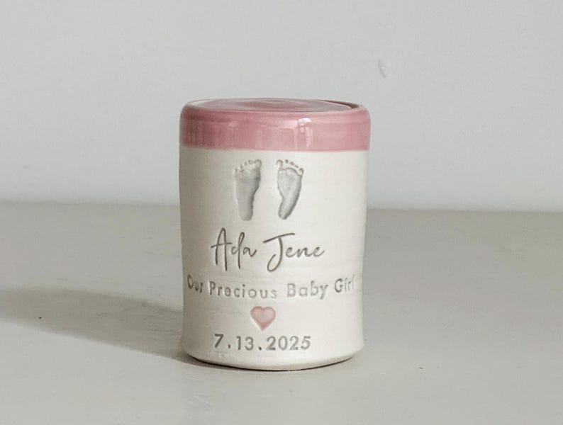 A small tall pink glaze and white porcelain keepsake infant urn. It features a custom footprint, the custom name, a phrase, a heart, and the date.