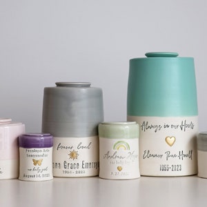 A collection of ceramic urns in various sizes and colors, including pink, gray, and teal. Some urns feature text such as names, dates, and the phrase "Always in our Hearts." The urns have a minimalist modern timeless design.