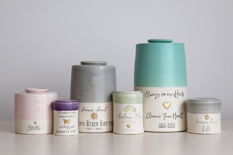A collection of ceramic urns in various sizes and colors, including pink, gray, and teal. Some urns feature text such as names, dates, and the phrase "Always in our Hearts." The urns have a minimalist modern timeless design.