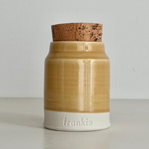 Image shows a collared shape urn with cork lid. It has an orange / wheat colored glazed upper section, and white unglazed clay lower portion. There is a custom name stamped into the clay